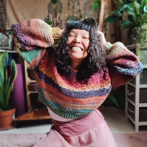 May include: A woman wearing a colorful striped crochet sweater with a pink skirt and lace trim. The sweater has a loose fit and long sleeves. The skirt is long and flowy.