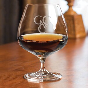 May include: A clear glass brandy snifter with a wide bowl and a long stem. The glass is filled with amber-colored brandy and has the initials "CNC" etched on the side.