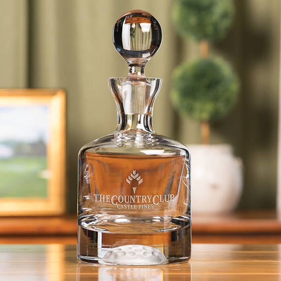 A hand-blown or crafted crystal decanter with an actual golf ball fused into the base — a genuinely surprising "wait, how did they do that?" piece of functional barware art.