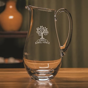 May include: A clear glass pitcher with a handle. The pitcher has an etched design of a tree with the text "WBGCC Est. 1918" underneath.