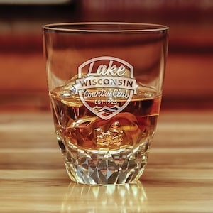 May include: A clear glass rocks glass with a diamond-shaped pattern. The glass is filled with amber liquid and ice cubes. The glass is etched with the words "Lake Wisconsin Country Club Est. 1925" in a shield shape.