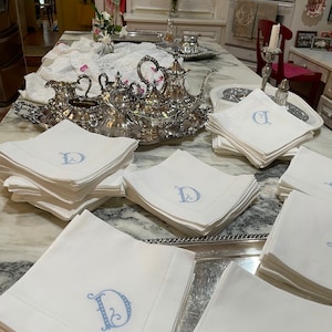 Puede incluir: Una mesa puesta con un juego de t&eacute; de plata y una pila de servilletas de lino blanco con letras D bordadas en azul.