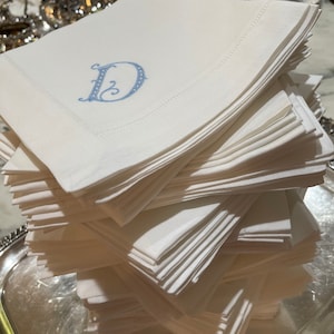 Puede incluir: Una pila de servilletas de lino blanco con una letra "D" bordada en azul en la servilleta de arriba.