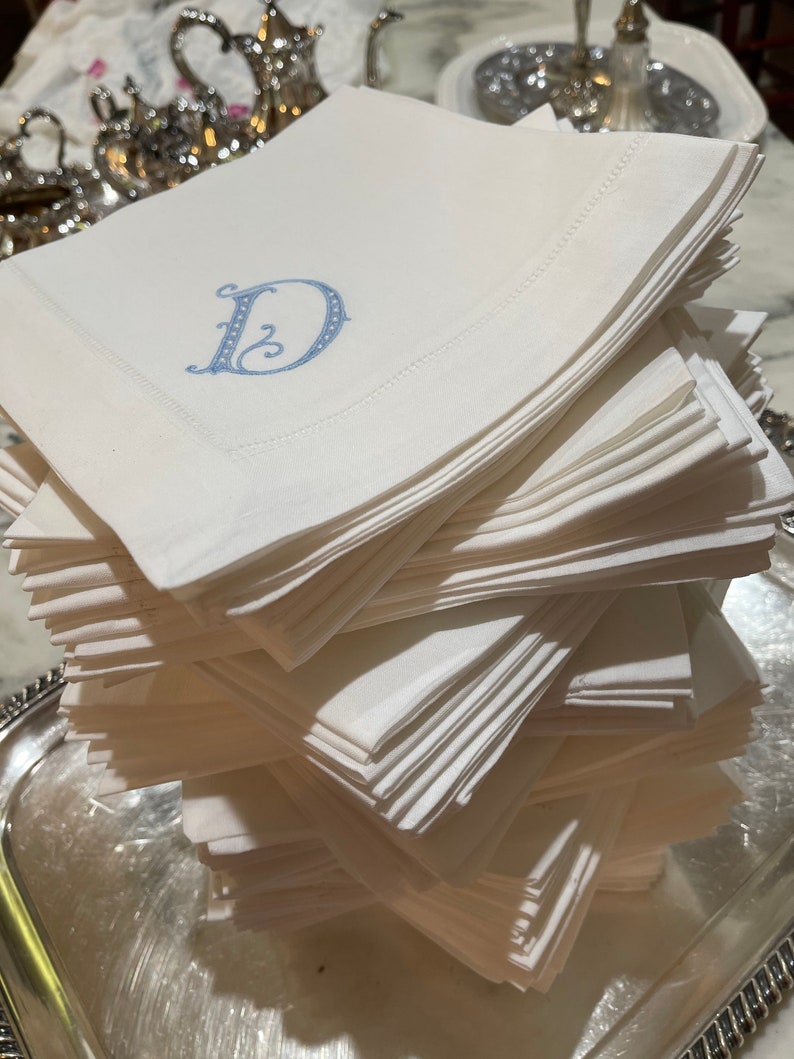 Puede incluir: Una pila de servilletas de lino blanco con una letra "D" bordada en azul en la servilleta de arriba.