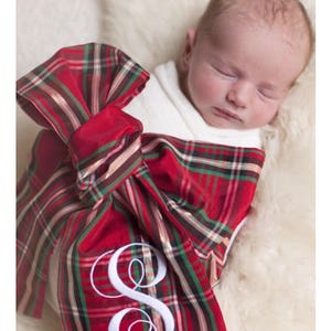 May include: A newborn baby swaddled in a red and green plaid fabric with a large bow and a white embroidered letter "S".