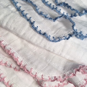 May include: A stack of white fabric baby burp cloths with pink and blue crocheted trim.
