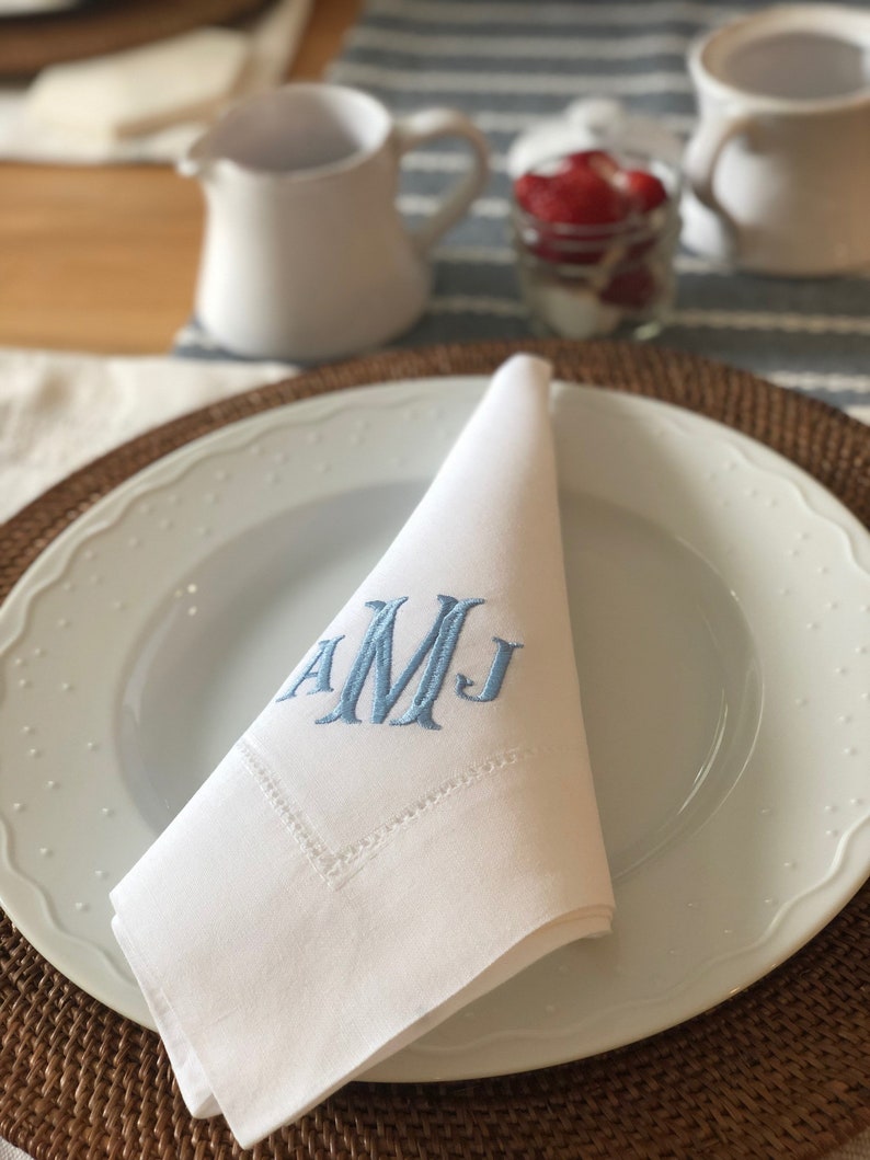 Puede incluir: Servilleta de lino blanco con iniciales bordadas en azul "AMJ" doblada en un plato blanco con un plato de mimbre marr&oacute;n.
