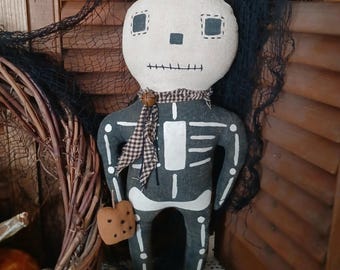 May include: A handmade Halloween skeleton doll with a cream-colored head and a gray body with white bone details. The doll has a checkered scarf and holds a heart-shaped cookie. A black net and a woven wreath are in the background.