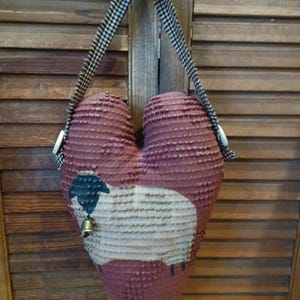 May include: A heart-shaped fabric ornament in a deep maroon color with a textured surface. A cream-colored sheep illustration with a black head and a small metal bell is centered on the front. It hangs from a black and white gingham ribbon.