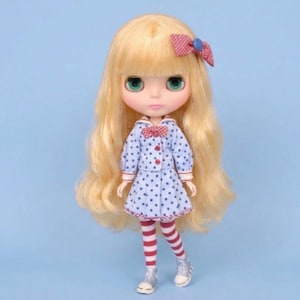 Pode incluir: Uma boneca com longos cabelos loiros ondulados e olhos verdes, vestindo um vestido de gola azul claro com bolinhas azul-escuras, um laço xadrez vermelho e branco no pescoço, acessório de cabelo a condizer, meias às riscas vermelhas e brancas e ténis prateados.