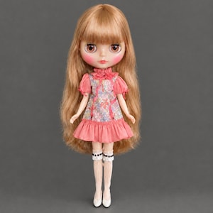 May include: A doll with long light brown hair and large brown eyes. The doll wears a short-sleeved dress featuring a floral pattern in shades of pink, blue, and green, accented with a ruffled collar and a pink skirt. White knee-high socks with black trim and white shoes complete the outfit.