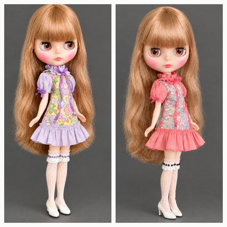 May include: Two dolls with long light brown hair and large eyes. The left doll wears a lavender dress with a floral pattern and purple ribbon, white ruffled socks, and white shoes. The right doll wears a coral dress with a floral pattern, white ruffled socks, and white shoes.