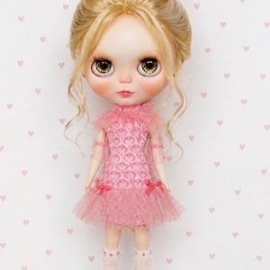 May include: A doll with blonde hair styled in a high bun, large brown eyes, and pale skin. The doll wears a pink sleeveless dress with a textured heart pattern, a ruffled tulle skirt, and small bows at the waist. It also wears white knee-high socks.