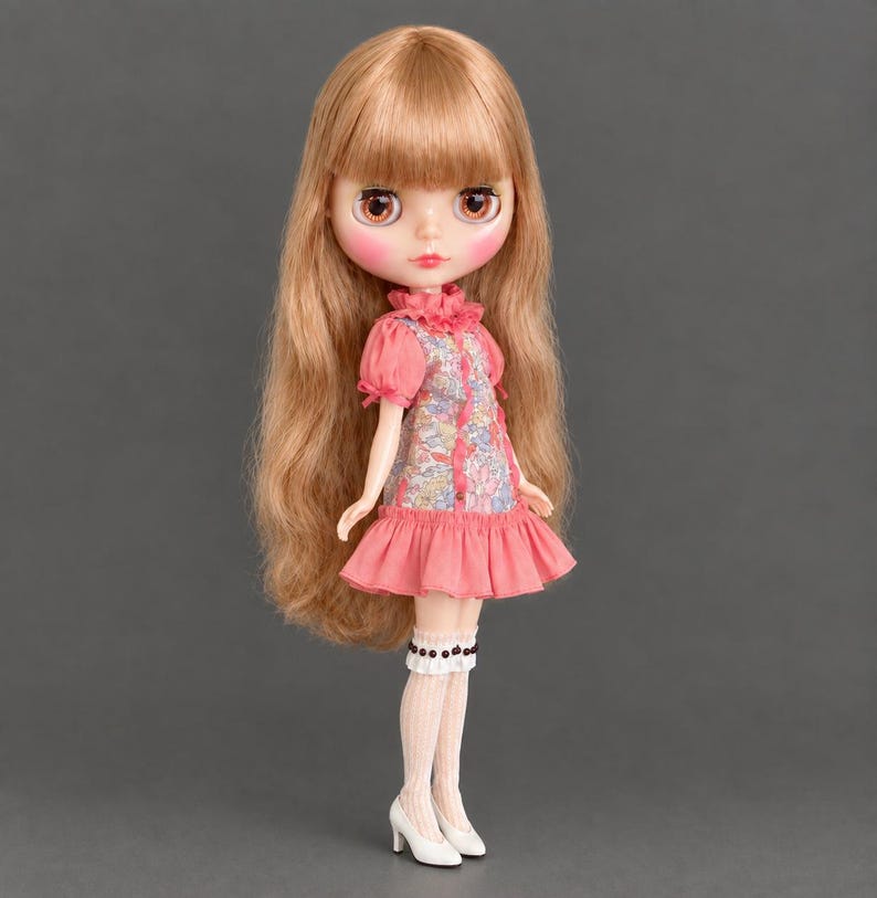 May include: A doll with long, wavy light brown hair, large brown eyes, and pink lips. The doll wears a short-sleeved dress with a floral bodice and a solid pink ruffled skirt, paired with white knee-high socks and white high-heeled shoes.