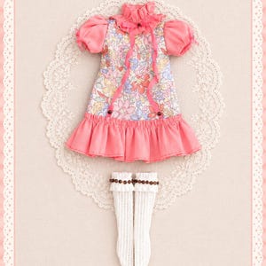 May include: A doll dress featuring a floral print bodice with pink puff sleeves, a ruffled collar with a thin pink ribbon, and a solid pink ruffled hem. The set includes a pair of white knee-high socks with a ribbed texture and brown bead trim.