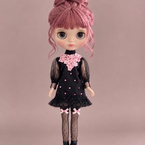 May include: A doll with dusty pink hair styled in a high bun with bangs. The doll wears a black dress featuring sheer puff sleeves, a pink floral collar, and pink sequins. The outfit includes black fishnet tights with pink bows and black boots.