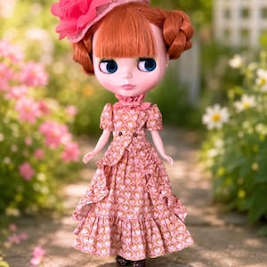 May include: A doll with ginger hair styled in two buns and bangs, wearing a pink hat with a large flower accent. The doll is dressed in a pink dress featuring a repeating heart pattern, a ruffled skirt, and a high collar with a small pink bow.