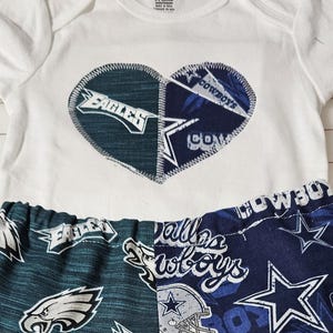 May include: A white short-sleeved bodysuit featuring a heart-shaped patch with Philadelphia Eagles and Dallas Cowboys logos, paired with matching split-leg pants. The pants display team logos and text on a teal and navy blue background over a ruler.