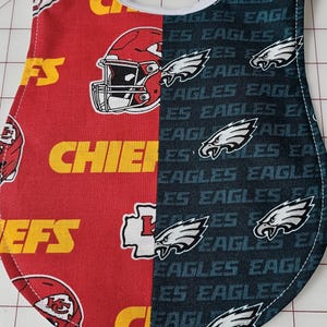 Pode incluir: Um babador para bebé vermelho e preto com os logotipos dos Kansas City Chiefs e dos Philadelphia Eagles. O babador é feito de tecido e tem uma alça branca.