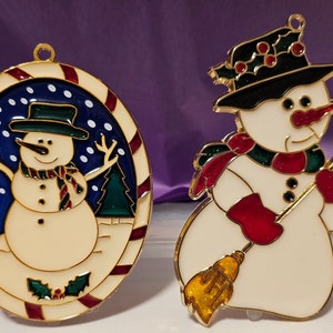 May include: Two stained glass snowman ornaments. One snowman is holding a broom and wearing a red and green scarf. The other snowman is in a blue and white winter scene with a candy cane border.