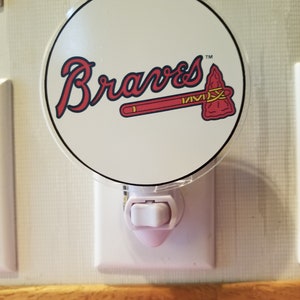 Pode incluir: Uma luz noturna branca com uma capa de plástico transparente que apresenta o logotipo dos Atlanta Braves. O logotipo é vermelho, azul e branco com a palavra "Braves" em uma fonte cursiva e um machado.