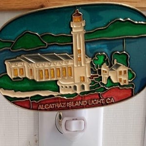 May include: Oval night light featuring a colorful depiction of Alcatraz Island Light, CA. The image shows a lighthouse, buildings, and surrounding landscape in shades of white, gold, green, blue, and red. The text "ALCATRAZ ISLAND LIGHT, CA" is visible.