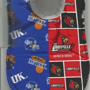 May include: A baby bib with a split design. One side is blue with "UK" and "Kentucky" text, a football helmet, and a basketball graphic. The other side is red with "Louisville Cardinals" text and a cardinal bird logo.