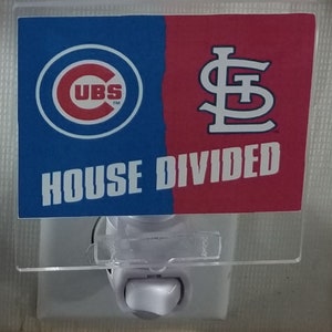 Pode incluir: Uma tampa de interruptor de luz de plástico branco com uma sobreposição de plástico transparente. A sobreposição tem um design vermelho e azul com os logotipos dos Chicago Cubs e dos St. Louis Cardinals. O texto "HOUSE DIVIDED" está impresso na sobreposição.