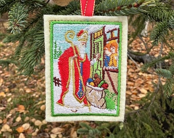 Puede incluir: Un adorno rectangular de fieltro con una escena bordada de San Nicolás con túnica roja y blanca y un báculo dorado, junto a un saco de regalos cerca de una puerta verde, acabado con una cinta roja para colgar.