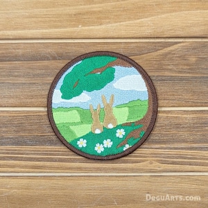 May include: Embroidered patch featuring two brown rabbits sitting under a green tree with white flowers. The background is a green field with a blue sky and white clouds.