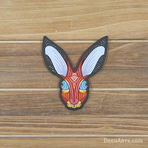 May include: A colorful, stylized patch featuring a rabbit design. The rabbit has long ears with white and blue inner ears, a red and orange face, and blue eyes. The patch is set against a wooden background.