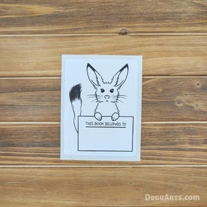 以下が含まれることがあります： 「THIS BOOK BELONGS TO」と書かれた看板を持つ、白黒の漫画の動物のイラストが描かれた白いステッカー。ステッカーは木製の表面に貼られています。