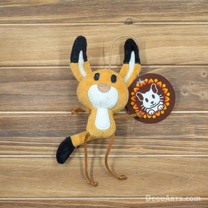 May include: A plush orange and black fox character with a white face and long, thin legs. The fox is holding a brown circle tag with a white cat logo and the text "DeguArts.com".