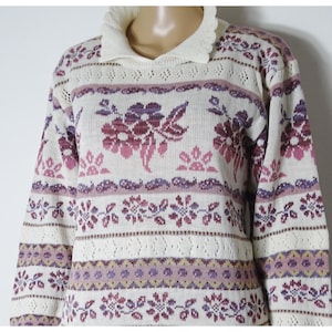 Könnte beinhalten: Ein weißer Strickpullover mit einem rosa und violetten Blumenmuster. Der Pullover hat einen Kragen und lange Ärmel.