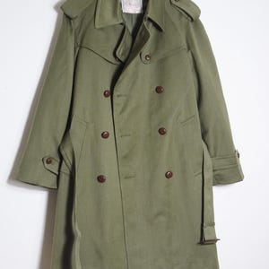 K&ouml;nnte beinhalten: Ein gr&uuml;ner, zweireihiger Trenchcoat mit braunem G&uuml;rtel und braunen Kn&ouml;pfen. Der Mantel hat einen Kragen und einen Sturmkragen.