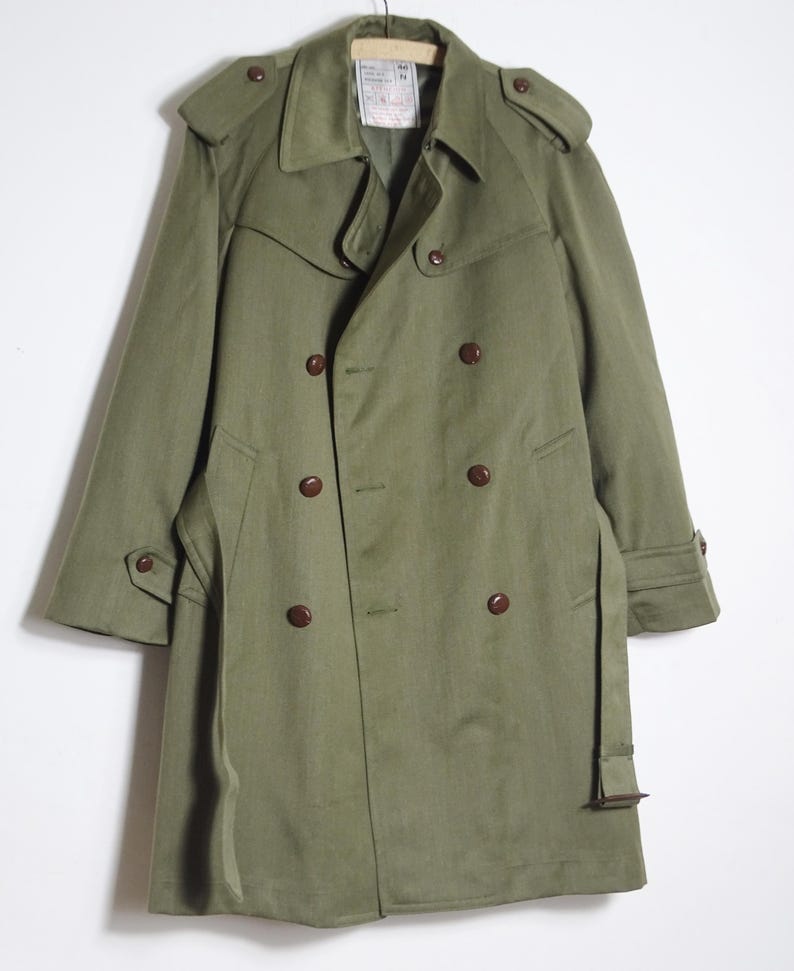 K&ouml;nnte beinhalten: Ein gr&uuml;ner, zweireihiger Trenchcoat mit braunem G&uuml;rtel und braunen Kn&ouml;pfen. Der Mantel hat einen Kragen und einen Sturmkragen.