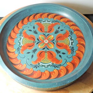 May include: A round, hand-painted wooden tray with a teal rim and a central design. The design features orange floral motifs and a teal background with accents of white, yellow, and black. The tray is likely used for serving or display.