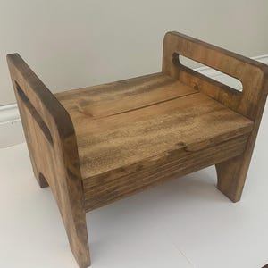 May include: A small wooden stool with a natural brown finish, featuring a rectangular seat and two side panels that extend upward to form integrated handles. The stool has four sturdy legs and a visible wood grain texture throughout the surface.