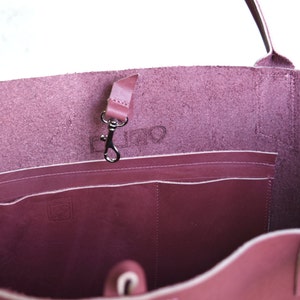 May include: The inside of a burgundy leather tote bag with a zippered pocket and a small leather strap with a metal hook. The bag has a white stitch detail around the edges.