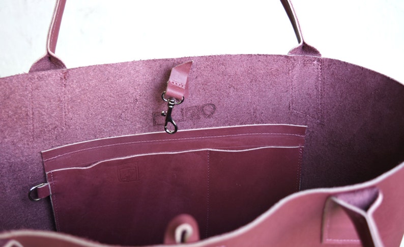 May include: The inside of a burgundy leather tote bag with a zippered pocket and a small leather strap with a metal hook. The bag has a white stitch detail around the edges.