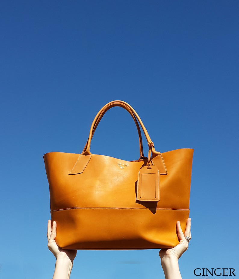 May include: A large, tan leather tote bag with a single strap and a small leather tag hanging from the strap. The bag is held up in the air against a bright blue sky.