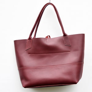May include: A burgundy leather tote bag with two top handles. The bag has a simple design with a single compartment and a small pocket on the front.