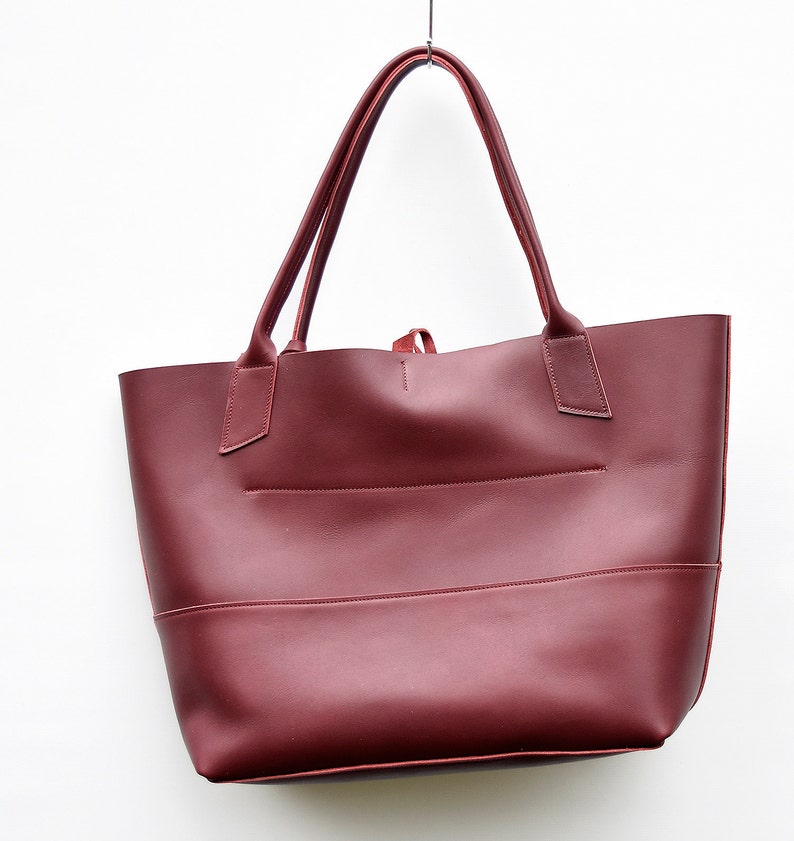 May include: A burgundy leather tote bag with two top handles. The bag has a simple design with a single compartment and a small pocket on the front.