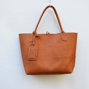 May include: A brown leather tote bag with a small leather tag hanging from the handle. The bag has a simple design and is made of high-quality leather.