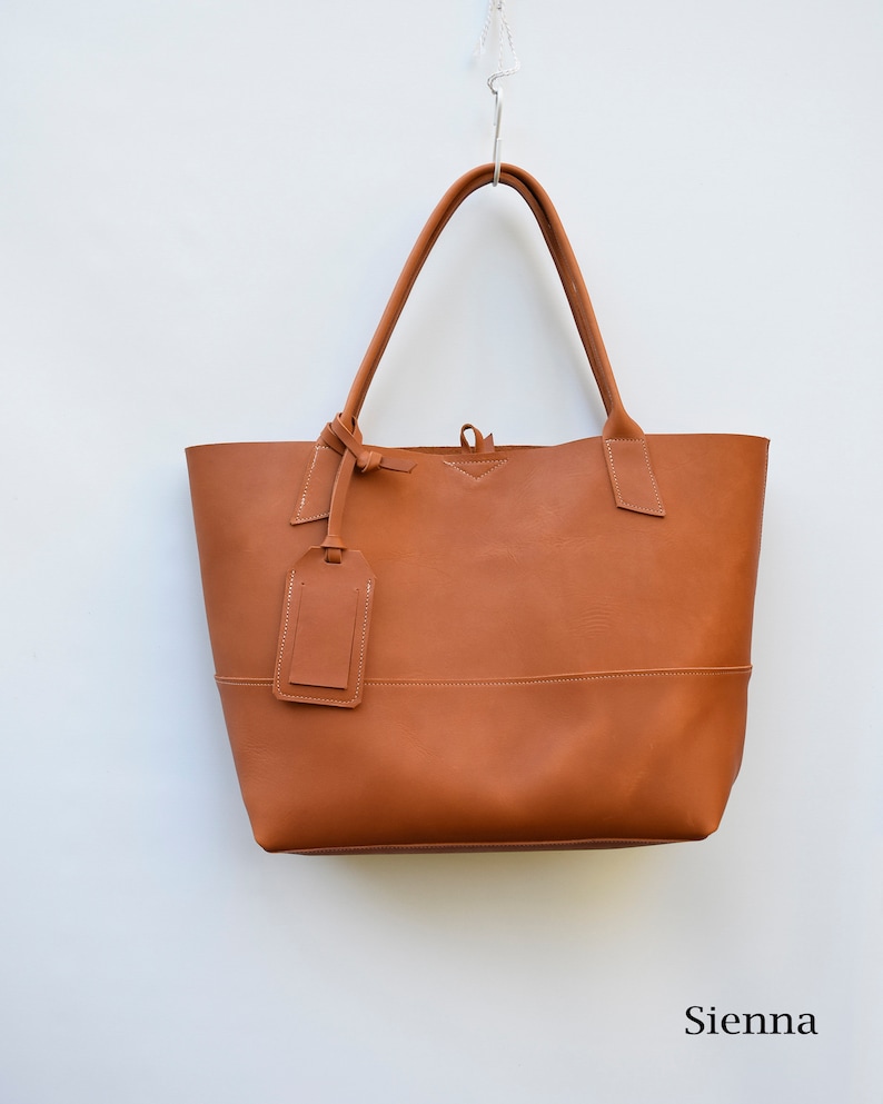 May include: A brown leather tote bag with a small leather tag hanging from the handle. The bag has a simple design and is made of high-quality leather.