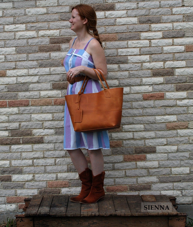 May include: A woman wearing a blue and white striped dress and brown leather boots is holding a brown leather tote bag. The bag has a brown leather strap and a brown leather tag. The woman is standing in front of a brick wall.