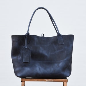 May include: A large, dark blue leather tote bag with a single, dark blue leather tag hanging from the front. The bag is sitting on a wooden stool.