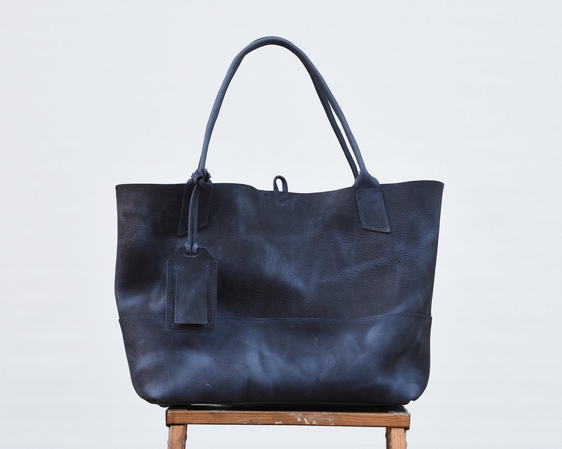 May include: A large, dark blue leather tote bag with a single, dark blue leather tag hanging from the front. The bag is sitting on a wooden stool.