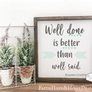 May include: A wooden sign with a white background and a blue and grey quote that reads "Well done is better than well said. - Benjamin Franklin". The sign is framed in a dark wood frame and is sitting on a table with two potted plants in front of it.