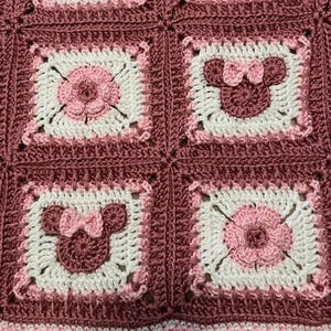 May include: A handmade crochet blanket featuring a repeating pattern of squares. Each square has a white center with either a pink flower or a Minnie Mouse design. The blanket is bordered in a darker pink.